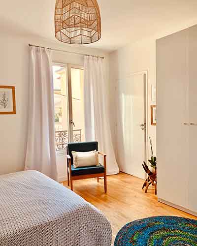 Chambre rénovée lumineuse avec parquet bois, murs clairs, fauteuil scandinave et décoration épurée.